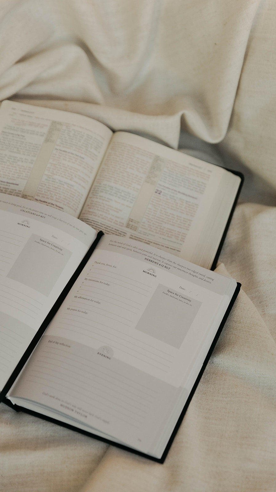 Open Journal with printed text pages laid on top of open bible  book with lined writing pages, set on a soft beige fabric background.