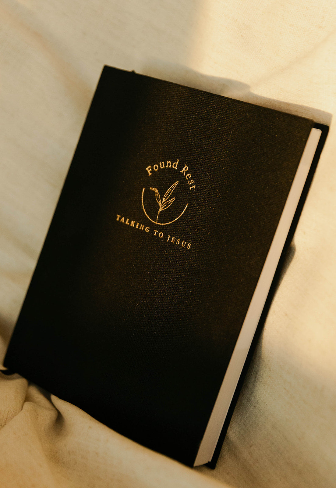 A black hardcover journal or Bible with gold foil lettering that reads 'Found Rest TALKING TO JESUS' and a wheat-like emblem, resting on a beige fabric surface.