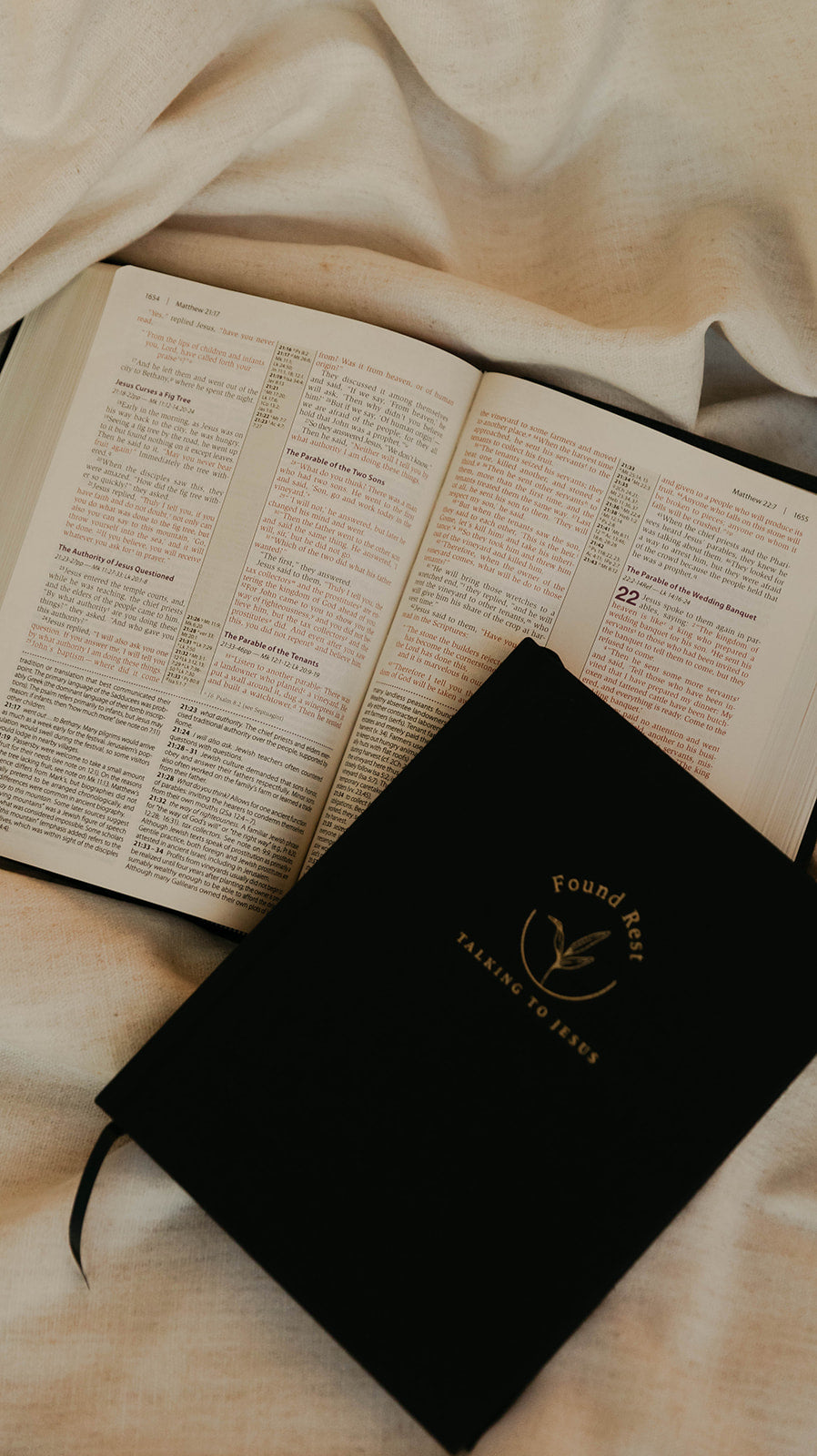 A black Bible study notebook with gold foil lettering lies on top of an open Bible, styled on draped cream-colored fabric.