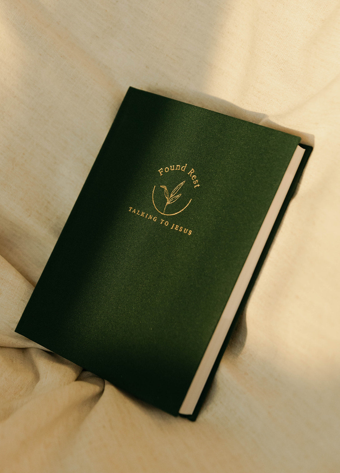 A dark green hardcover journal with gold foil branding reading 'Found Faith Talking to Jesus' and a small leaf emblem, placed on a beige fabric surface.