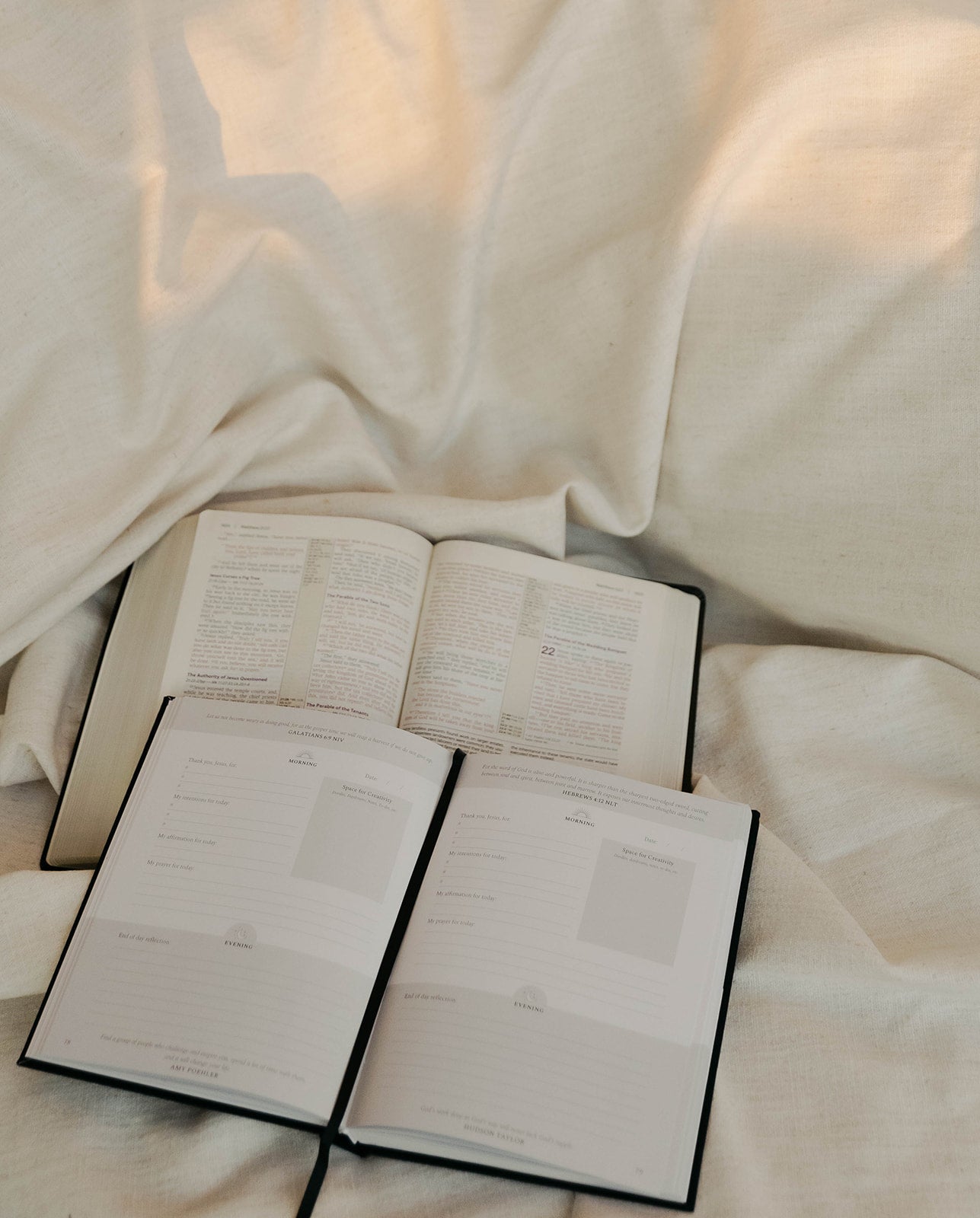 An open book with printed pages lies on draped white fabric, with two additional open booklets or notebooks placed in front of it.