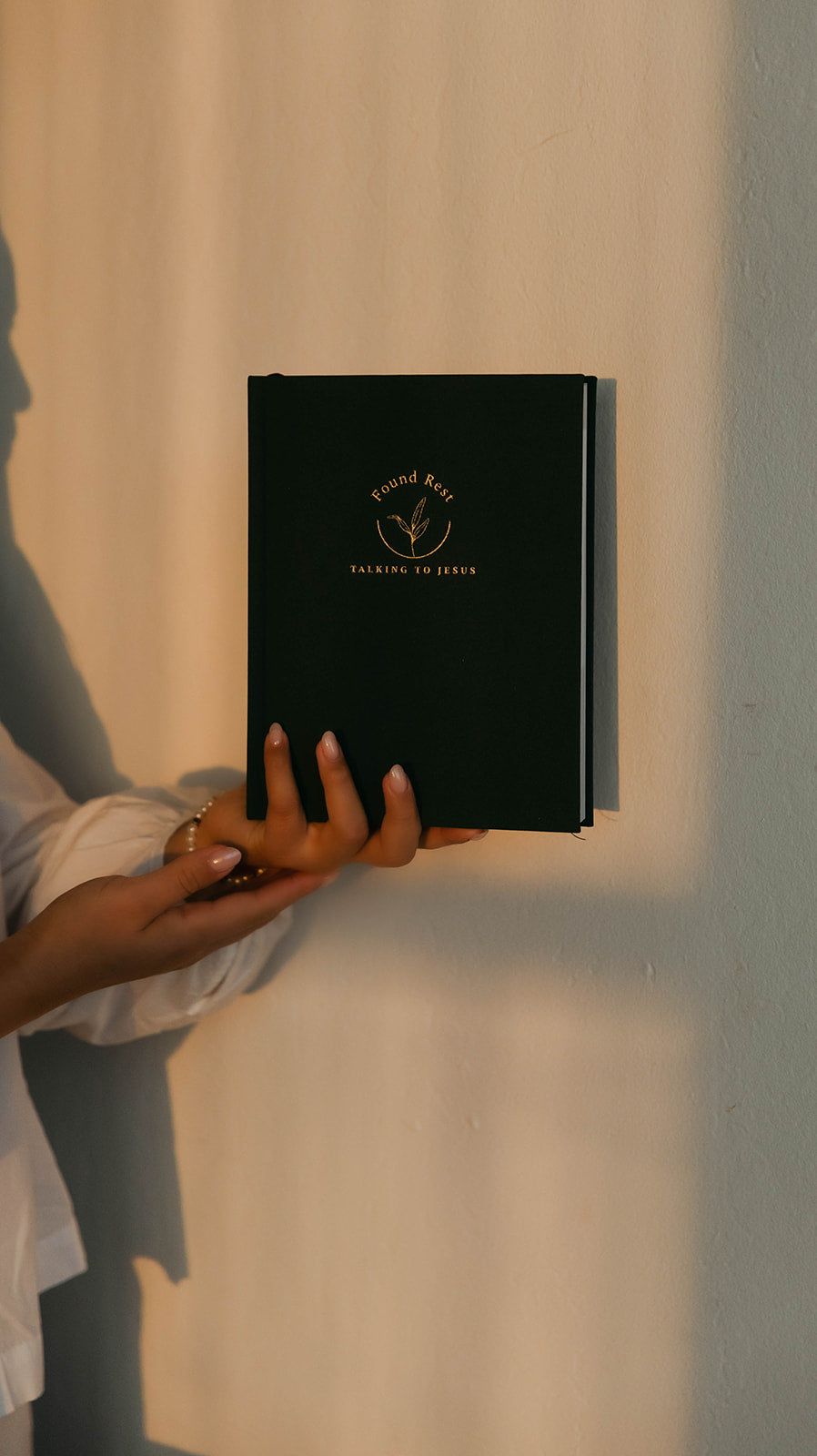 A person in a white shirt holds a slim black notebook or journal with gold foil branding against a warm beige wall with strong sunlight and shadows.
