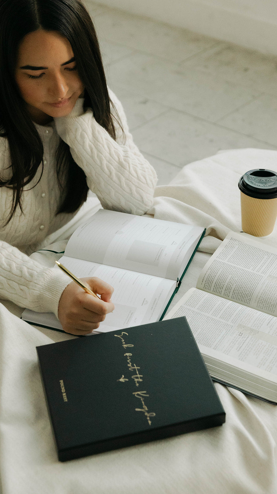 A woman in a white knit sweater sits on a bed writing in an open notebook, with books and a takeaway coffee cup nearby. A black book with gold script is prominently placed in the foreground.