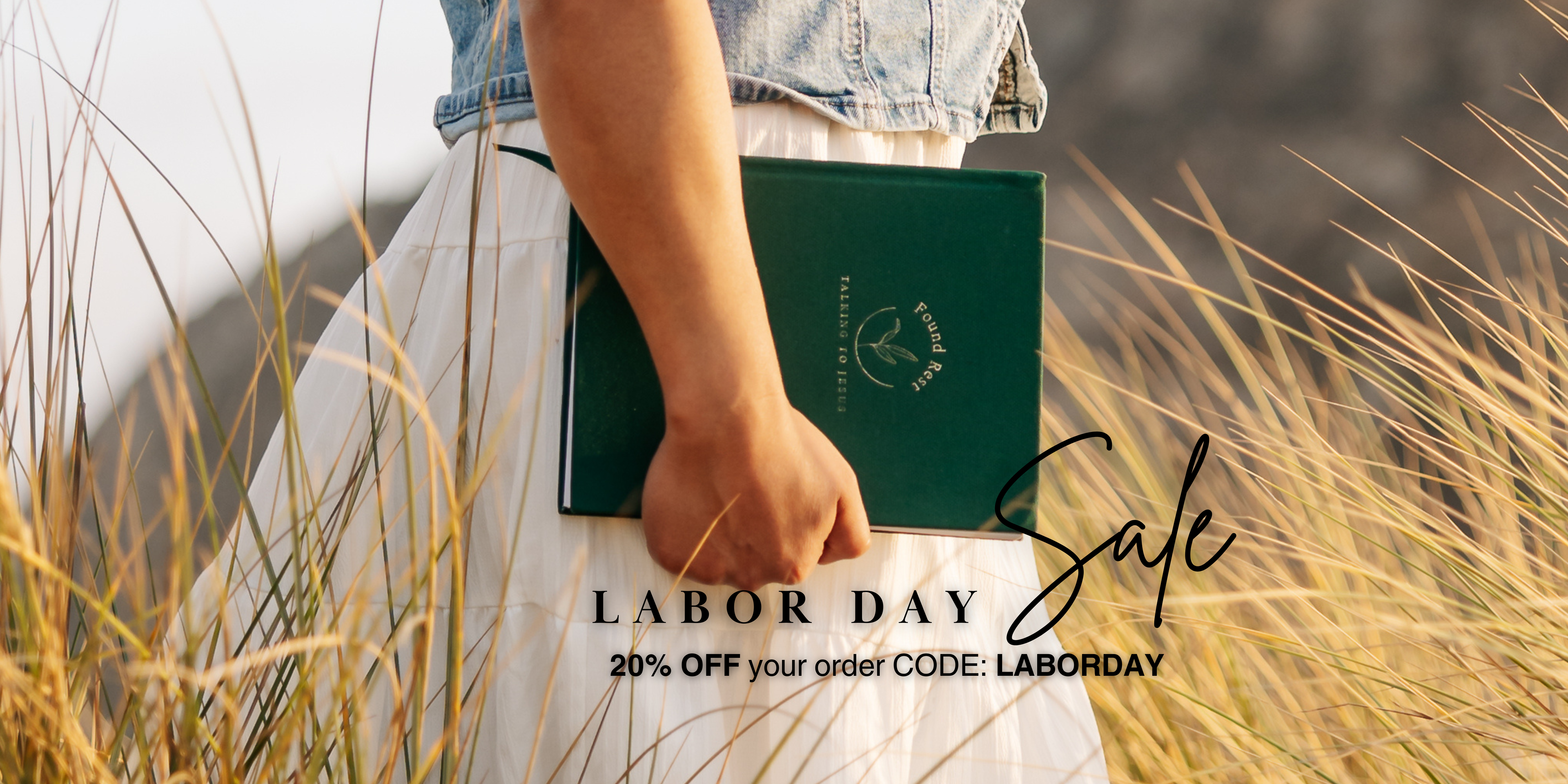 A person in a white dress and denim top holds a dark green book or folder with a gold logo in a field of dry grass. Text overlay reads 'LABOR DAY Sale 20% OFF your order CODE: LABORDAY'.